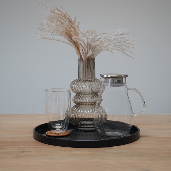 Zwart rond dienblad met een glazen karaf, glas, geribbelde vaas met siergrassen op een houten tafel.