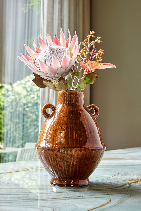 Bruine keramische vaas met twee handvatten, gevuld met een grote roze protea en takken.