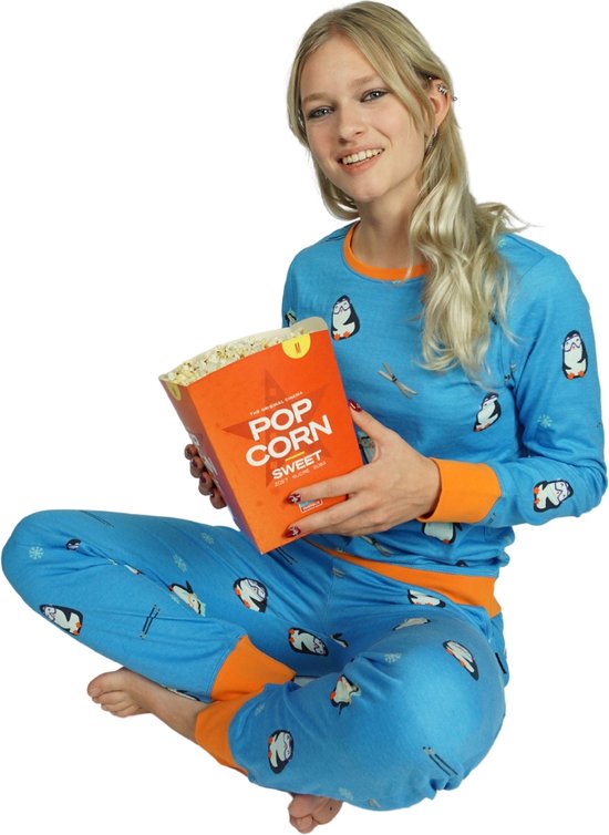 Model zittend in blauw pyjamapak met pinguïnprint en oranje boorden, met een doos zoete popcorn.