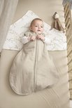 Baby in zandkleurige, gevoerde slaapzak met rits. Ligt op een laken met takjesprint in een ledikant.