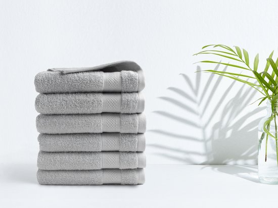 Stapel lichtgrijze handdoeken met een zachte textuur, tegen een witte achtergrond met een schaduw van een plant.