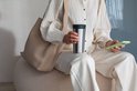 Vrouw in een beige outfit zit op een poef en houdt een roestvrijstalen beker vast met een grijs deksel.