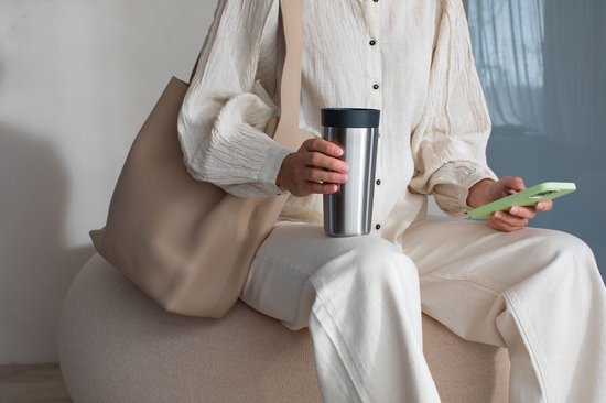 Vrouw in een beige outfit zit op een poef en houdt een roestvrijstalen beker vast met een grijs deksel.