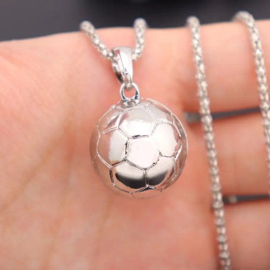 Hand die een zilverkleurige ketting met voetbalhanger vasthoudt. De hanger is van metaal met zeshoekige patronen.