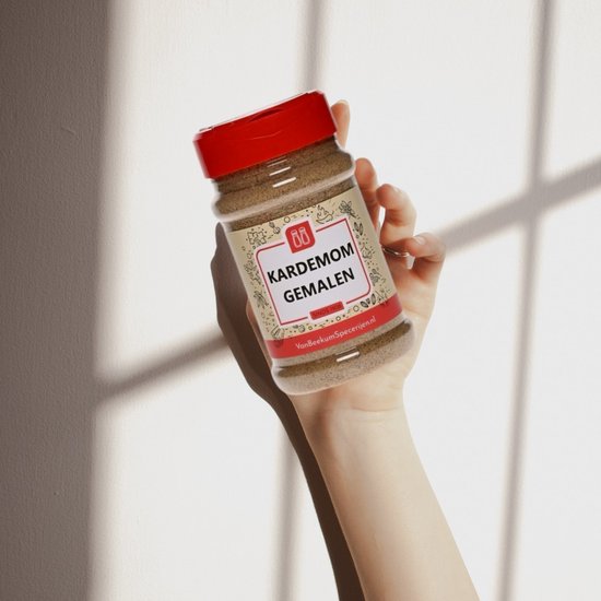 Hand met een rood-deksel kruidenstrooier met gemalen kardemom, etiket 'KARDEMOM GEMALEN'.