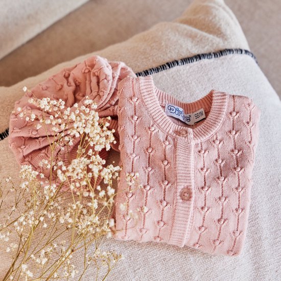 Roze gebreid vestje met hartjespatroon en knoopsluiting, naast een gevouwen mutsje.