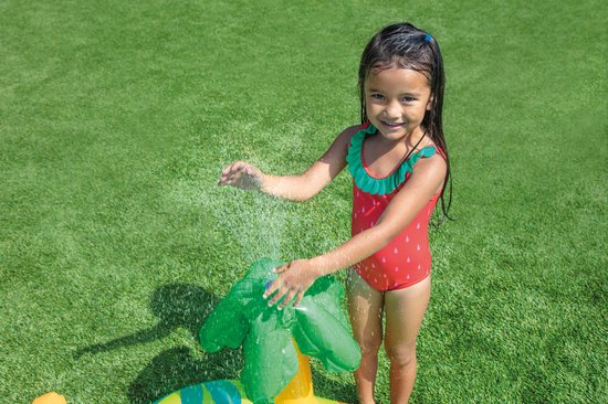 Een jong meisje in een rood badpak met aardbeienprint sproeit water op een groene dinosaurus waterspeeltje.