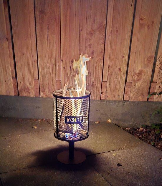 Brandende zwarte metalen staande vuurkorf met VOLT! logo, op een terras bij een houten schutting.