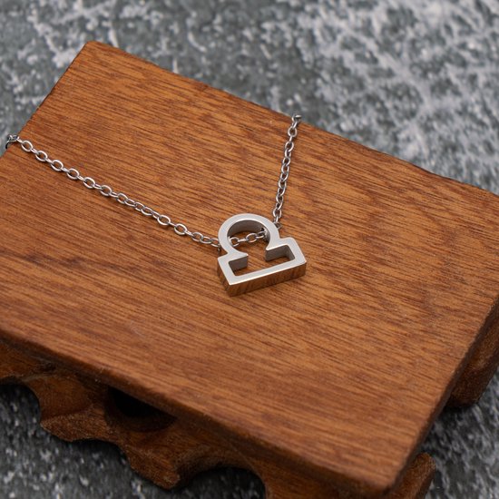 Collier argenté avec symbole Balance (Libra), posé sur un support en bois.