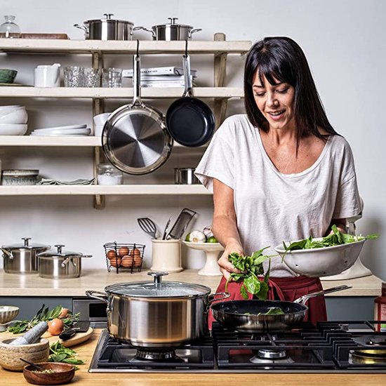 Een vrouw met lang zwart haar kookt met een KitchenAid koekenpan en grote pan, omringd door keukengerei en verse ingrediënten.