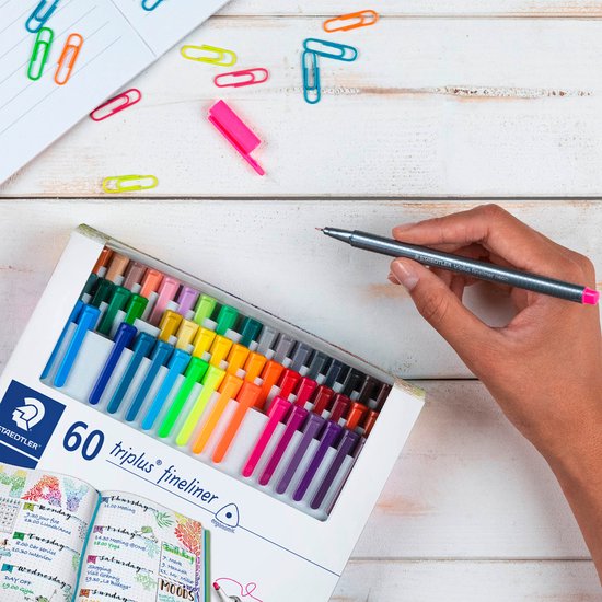 Main tenant un feutre Staedtler, au-dessus d'une boîte ouverte de 60 feutres de diverses couleurs.