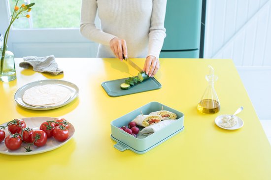 Une femme prépare un déjeuner avec des wraps, des radis et des tomates dans une boîte à pain Brabantia vert jade, sur un plan de travail jaune.