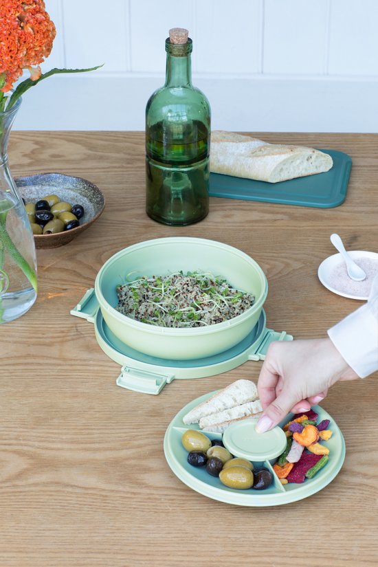 Boîte à salade vert pâle sur une table, avec quinoa, une assiette de légumes et d'olives.