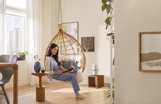 Une femme lit un livre dans un fauteuil suspendu dans un salon lumineux. À côté d'elle se trouve un humidificateur d'air Philips blanc.