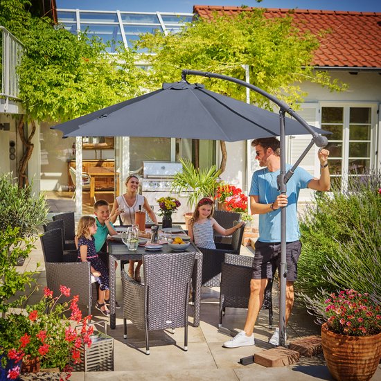 Een gezin geniet van een maaltijd aan tafel onder een donkergrijze zweefparasol op een zonnig terras.