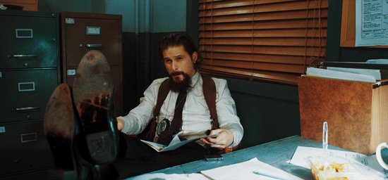 Een man met een baard en stropdas zit met de benen omhoog aan een bureau.