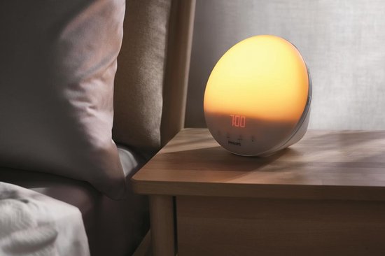 Een Philips wekker met oranje gloed staat op een houten nachtkastje naast een bed.