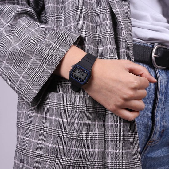 Un poignet portant une montre Casio F-91W-1Yef noire, portée avec un blazer à carreaux et un jean. L'écran indique 9:28.6.