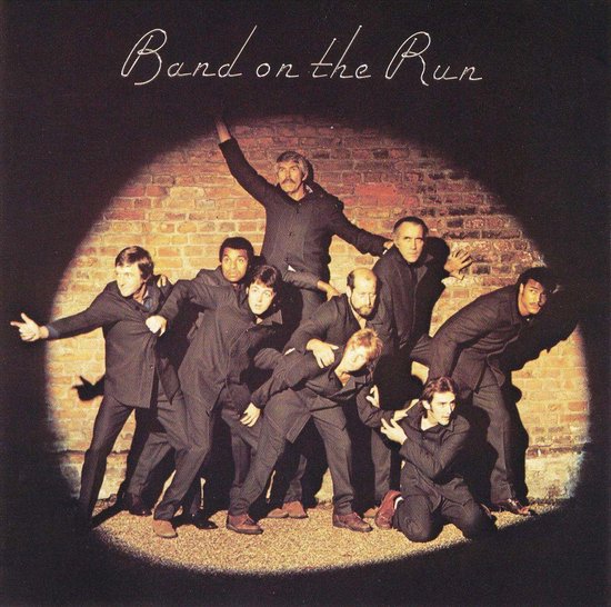 Een groep van negen mannen in donkere pakken, verlicht door een spotlight voor een bakstenen muur. De titel "Band on the Run" staat erboven.