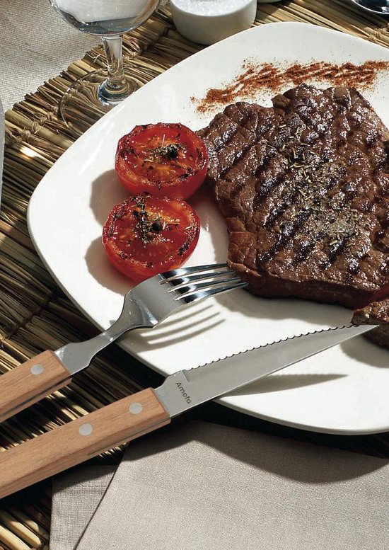 Een steakmes met kartelrand en houten handvat, naast een vork en gegrilde biefstuk met tomaten.