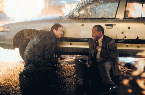 Twee mannen, beiden politieagenten, zitten op de grond in de regen bij een kogel-beschadigde auto.