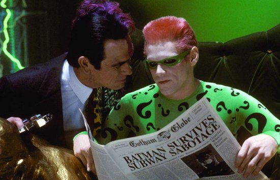 Two-Face kijkt naar de Riddler, die een krant vasthoudt met de kop 'Gotham Globe'. De Riddler draagt een groen pak met vraagtekens.