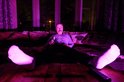 Man met snor zit op bank met verlichting in paars en roze, kijkt tv met drankje.
