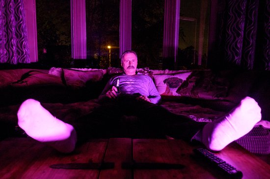 Man met snor zit op bank met verlichting in paars en roze, kijkt tv met drankje.