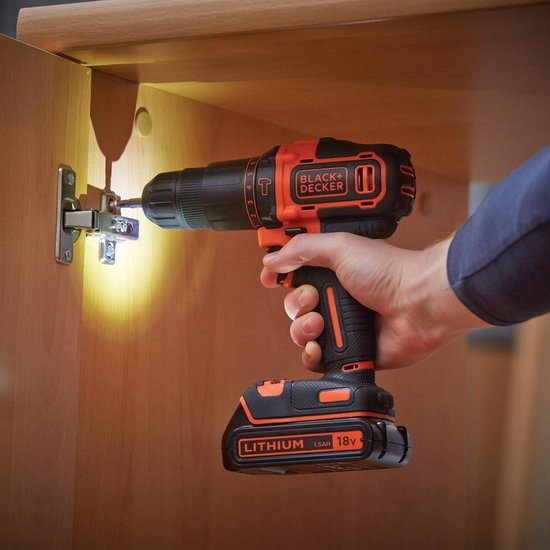 Hand met zwarte en oranje accuboormachine schroeft in een scharnier van een houten kastje.