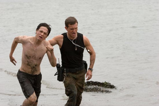 Twee mannen rennen door ondiep water. De linker man is ontbloot bovenlijf, de rechter man draagt een zwart tanktop.