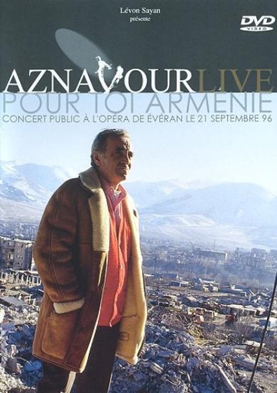 Charles Aznavour in een bruine lammy coat, voor een stadslandschap.