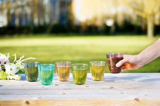 Un homme prend un verre brun-rouge sur une table en bois à l'extérieur. Cinq autres verres de couleurs différentes sont alignés.
