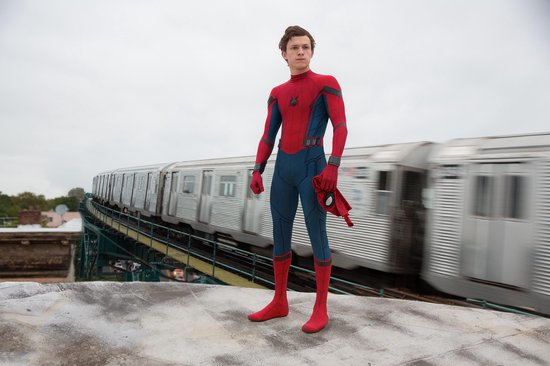 Een man in een rood en blauw Spiderman-pak staat op een dak, met een trein die op de achtergrond voorbij raast. Hij houdt een rood masker vast.