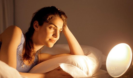 Vrouw kijkt uit het raam naast een Philips wekker die een zonsopkomst simuleert.
