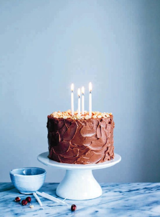 Chocoladetaart met brandende kaarsen op een witte taartschotel, naast een blauw kommetje.