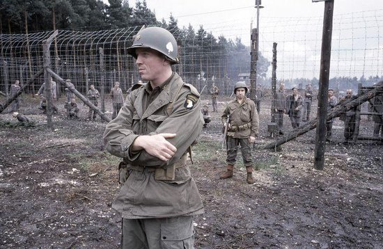 Soldaat in helm en uniform voor een omheind terrein met andere soldaten, met bomen op de achtergrond.