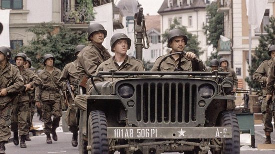 Soldaten in uniformen en helmen rijden in een open jeep, omringd door andere marcherende soldaten.