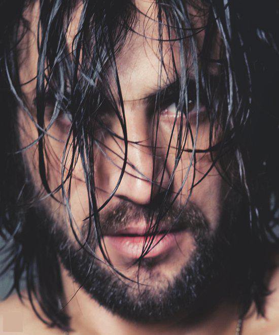 Man met nat zwart haar dat over zijn gezicht valt, met baard en intense blik.