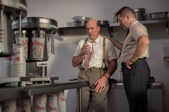 Twee mannen in een keuken, een met een bekertje McDonald's, de ander met hand op schouder. Veel McDonald's bekers op de voorgrond.