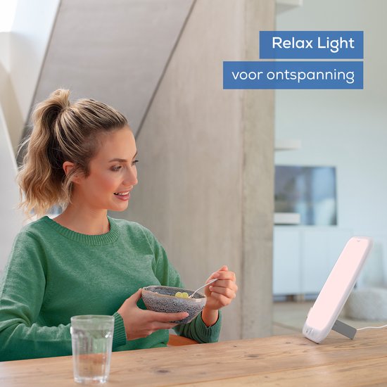 Vrouw bij tafel met Beurer daglichtlamp, 'Relax Light' voor ontspanning, eet uit kom.
