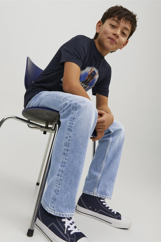 Jongen zittend op een stoel in een lichtblauwe spijkerbroek, donker T-shirt met opdruk en blauw-witte sneakers.