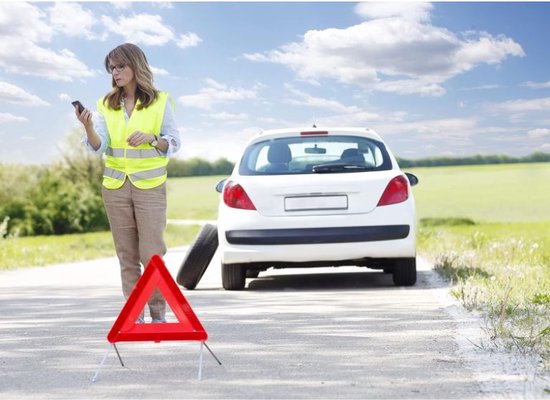 Vrouw in geel veiligheidsvest belt met telefoon bij een witte auto met lekke band. Gevarendriehoek op de weg.