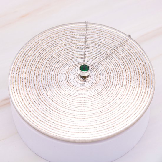 Zilverkleurige ketting met groene zirkonia hanger op een zilveren ronde ondergrond met patronen.