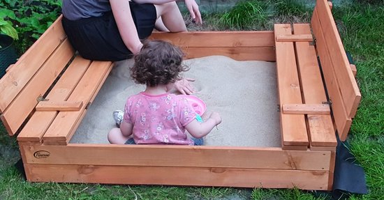 Een houten zandbak met inklapbare banken, met een jong kind zittend in het zand. De zandbak is van hout.