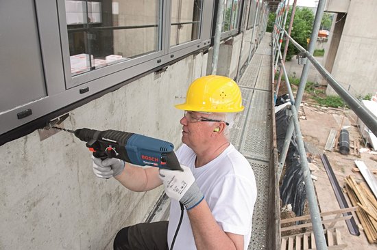 Man met gele helm gebruikt blauwe Bosch boorhamer om in betonnen muur te boren.