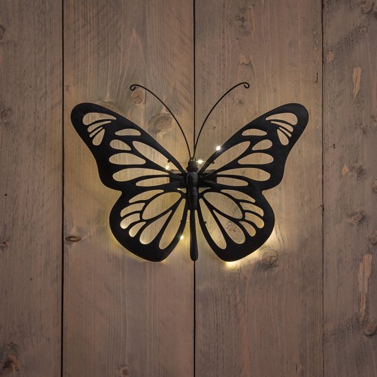 Une décoration papillon en métal noir aux ailes ouvertes, avec de petites lumières à l'arrière, sur un mur en bois.