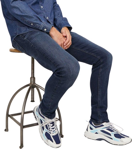 Man zittend op een kruk, draagt spijkerbroek, denim overhemd en sneakers.