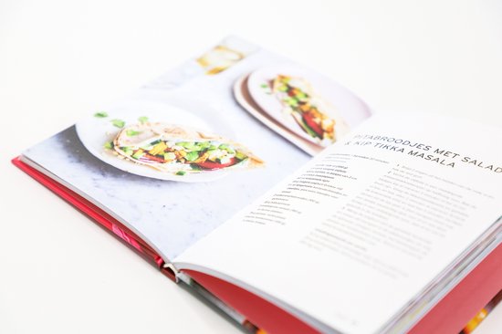 Open kookboek met recepten en een pita broodje gevuld met salade en kip, op een wit bord.