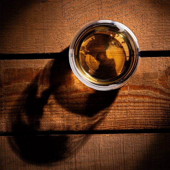 Bovenaanzicht van een glas gevuld met amberkleurige drank, erop de projectie van de wereldkaart, staand op een houten ondergrond.