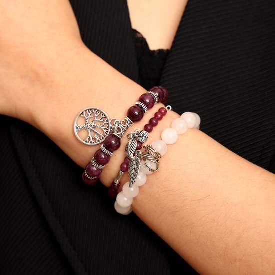 Drie gestapelde armbanden om een arm, met paarse en witte kralen en zilveren bedeltjes van een boom en bladeren.